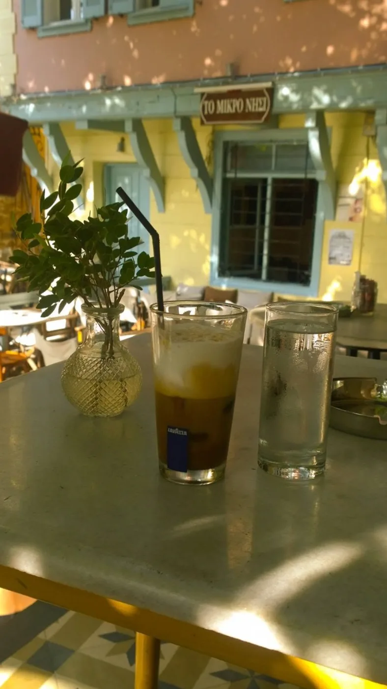 Ein Freddo Cappuccino und ein Glas Wasser auf einem Cafétisch im lichten Schatten